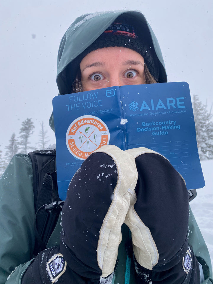 Wide-eyed female wearing big mittens holds AIARE blue book in front of her face during avalanche course.