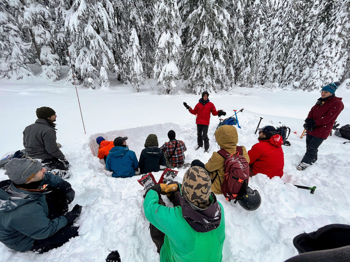 Students sit in the snow and listen to their avalanche course instructor discuss safety in the backcountry.