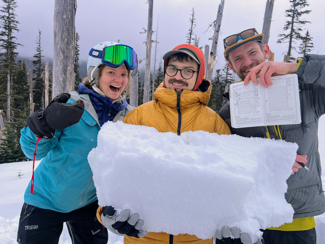 Three smiling AIARE students pose holding a large block of snow and decision-making blue book.