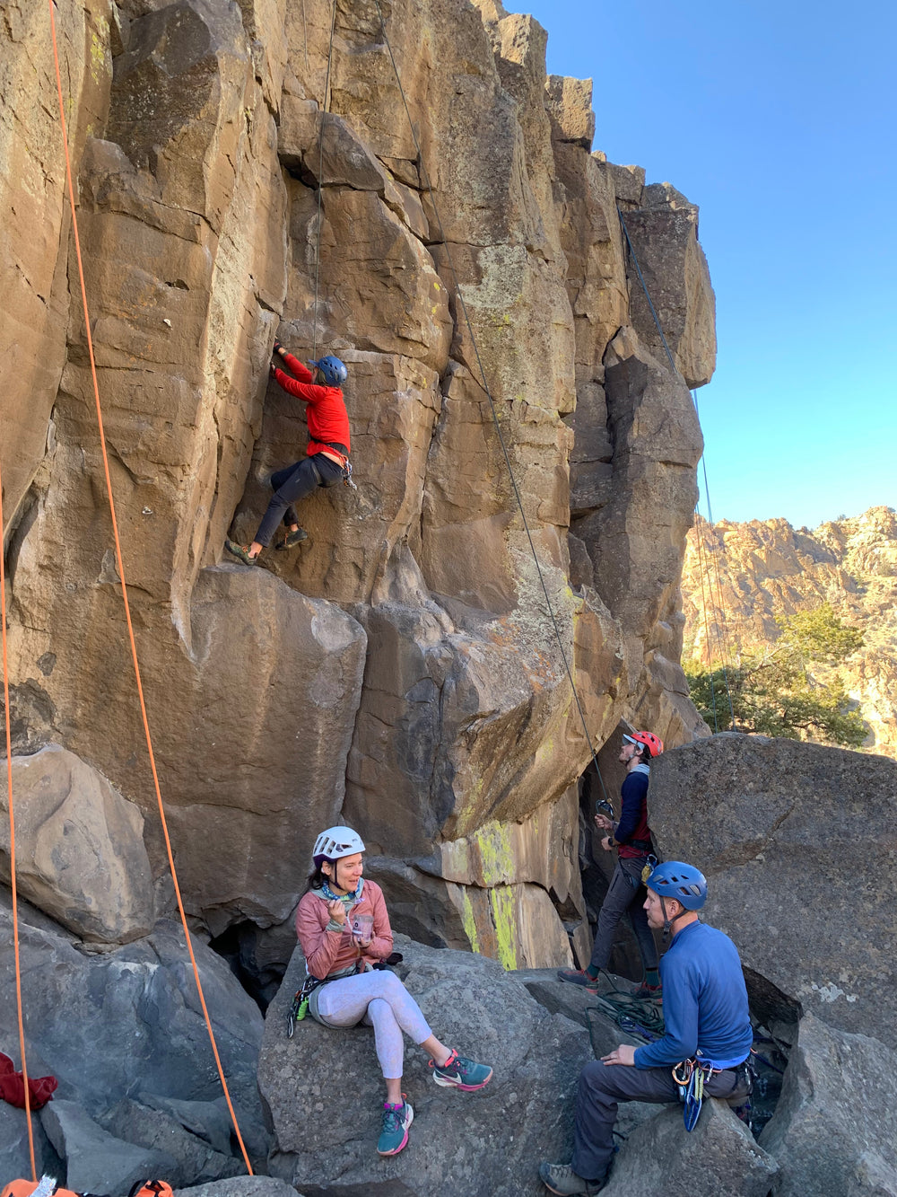 One climber is belaying another on a crack climb while two more converse on rocks in the foreground.